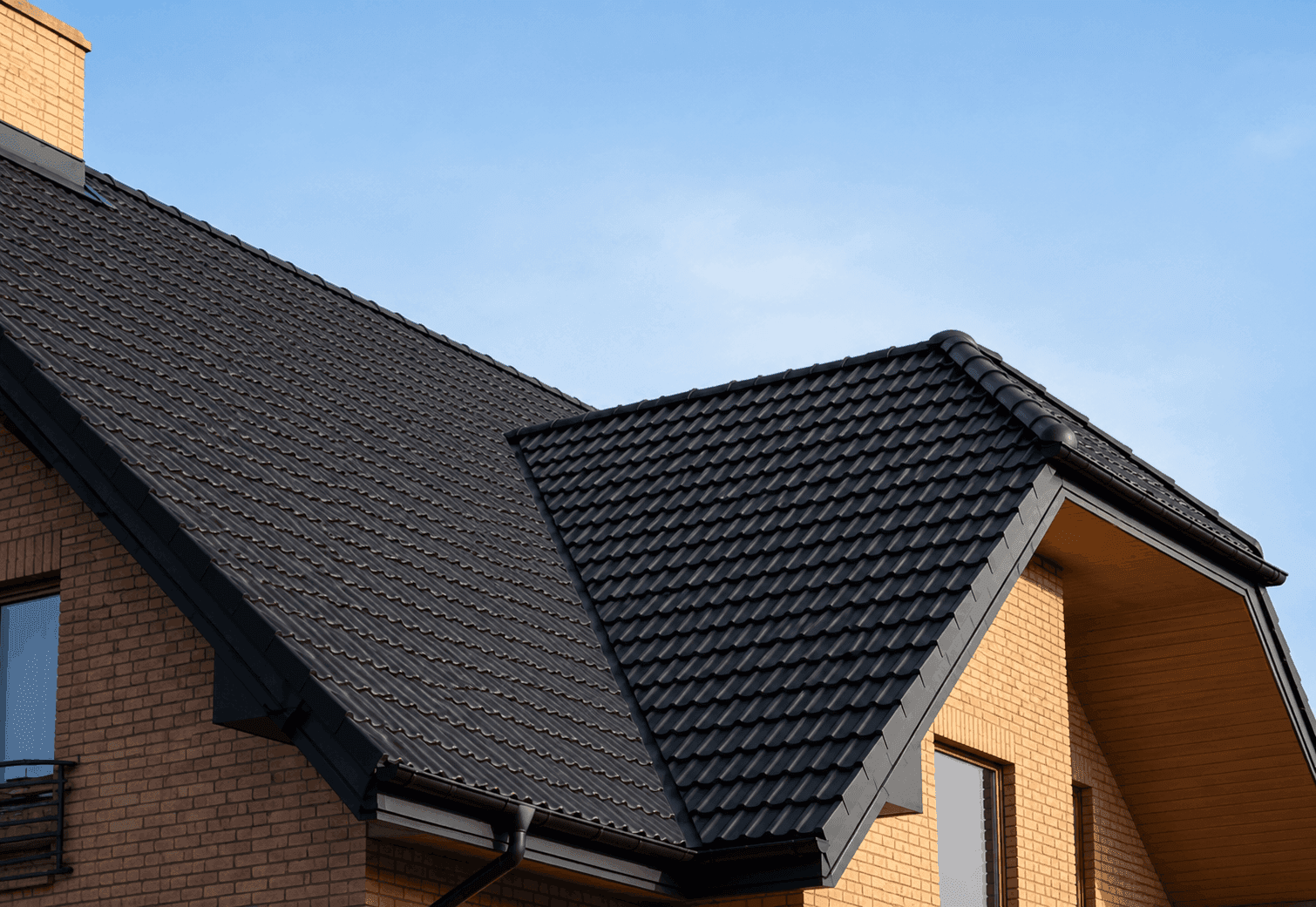 Close-up of a home roofline with dark tile roofing, gutters, and brick walls